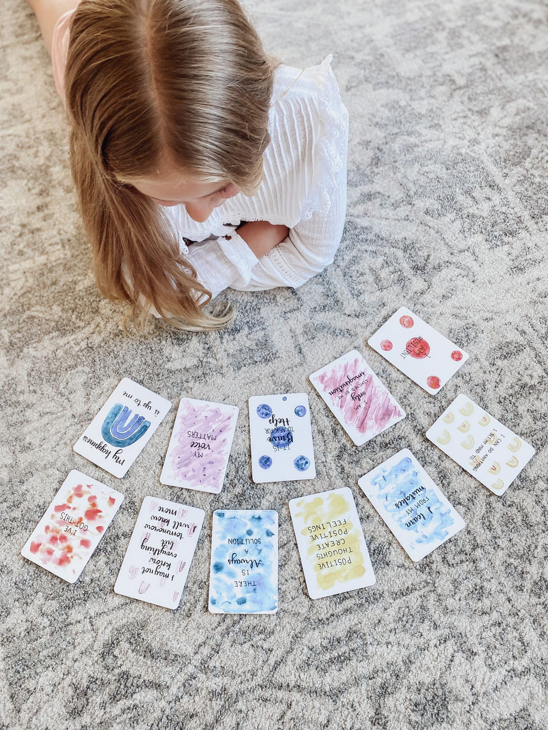 Young girl happily lying on the floor, engaged in exploring colorful, uplifting affirmation cards, promoting mental health and emotional resilience in children.